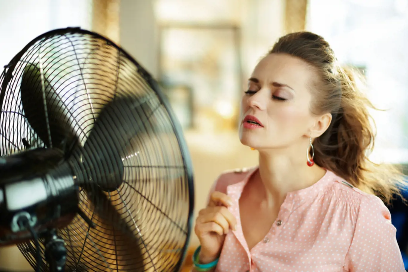 young housewife in the modern house in sunny hot summer day enjoying freshness in the front of working fan.