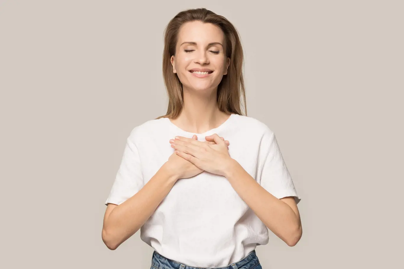 Happy peaceful millennial woman in casual clothes isolated on grey studio background hold hands at chest close to heart, smiling grateful female stand feel thankful, show appreciation and love