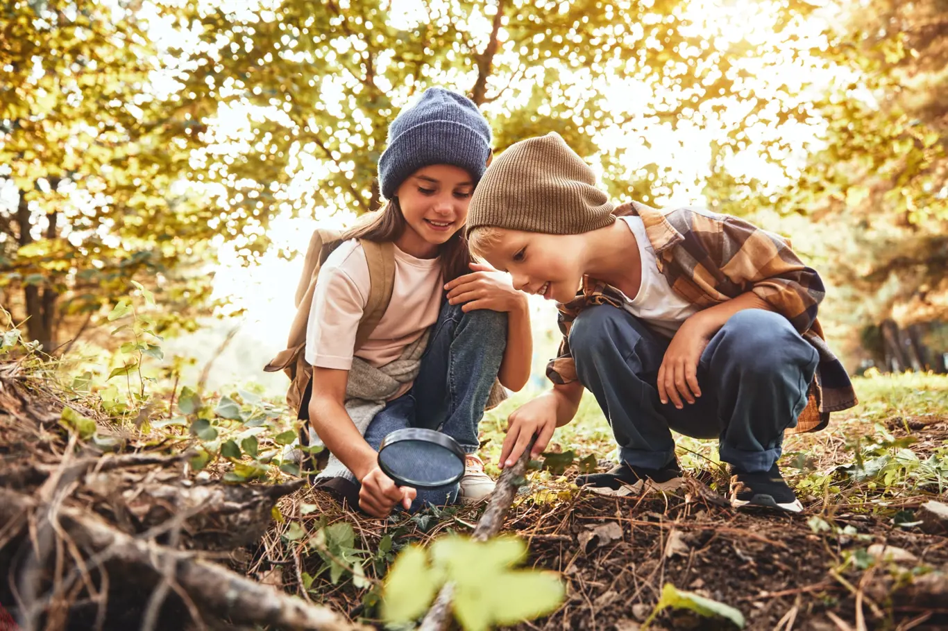 Two children boy and girl in warm hats with backpacks looking examining tree bark through magnifying glass while exploring forest nature and environment on sunny day during outdoor ecology school less