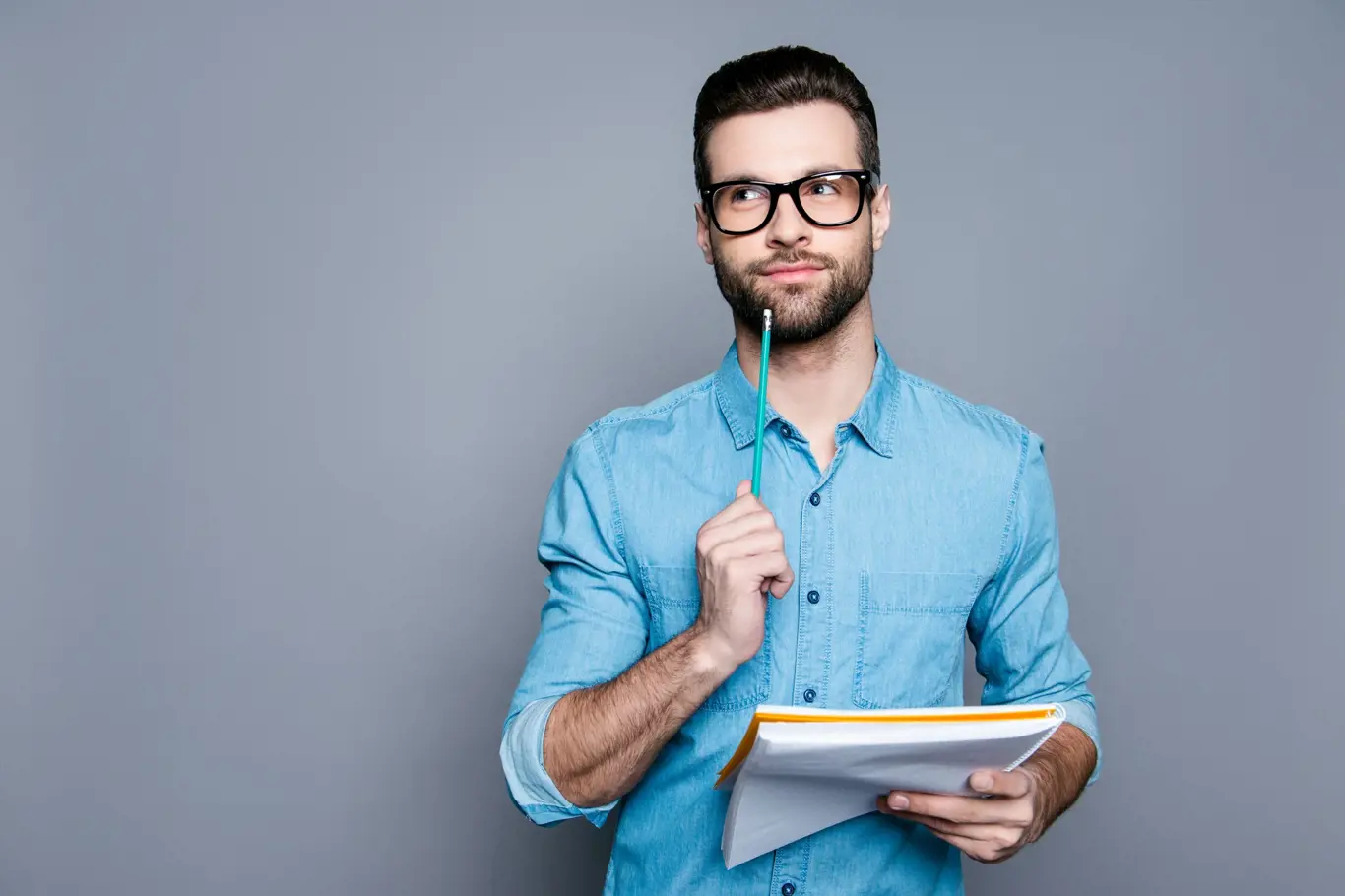 Young bearded guy in glasses holding a notebook and thinking about exams.