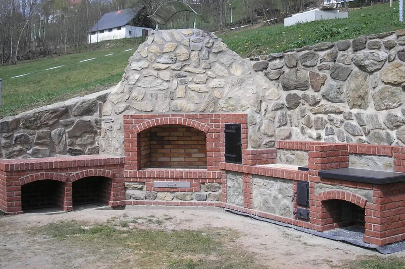 Velkorysá rustikální venkovní kuchyně s otevřeným ohništěm skrývá výkonné moderní topidlo a dostatek místa na uložení dřeva i přípravu pokrmů. 