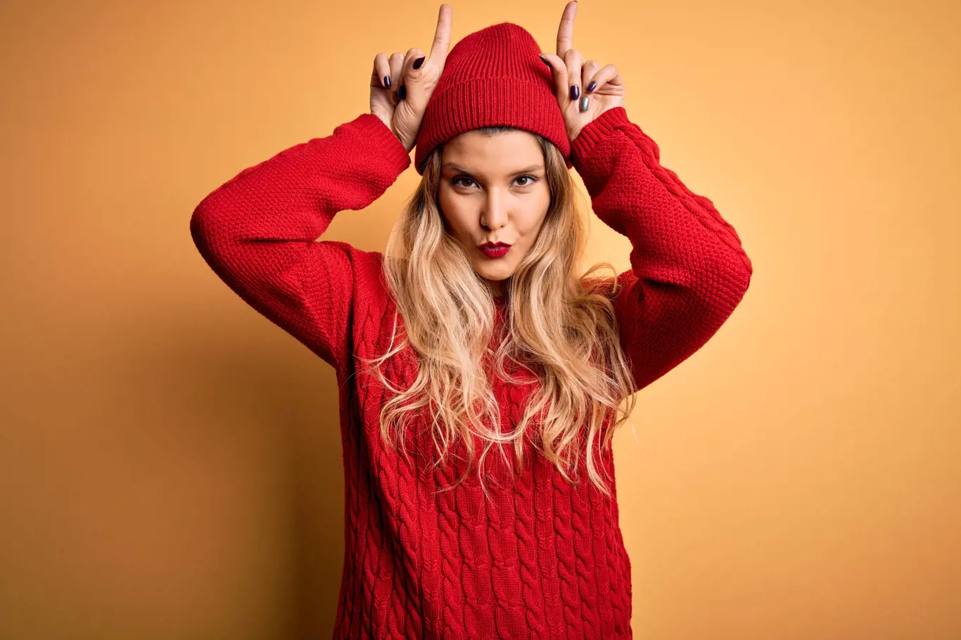 Young beautiful blonde woman wearing casual sweater and wool cap over white background doing funny gesture with finger over head as bull horns