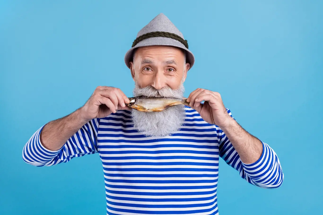 Portrait of attractive hungry cheerful grey-haired man biting fresh fish food carp isolated over bright blue color background