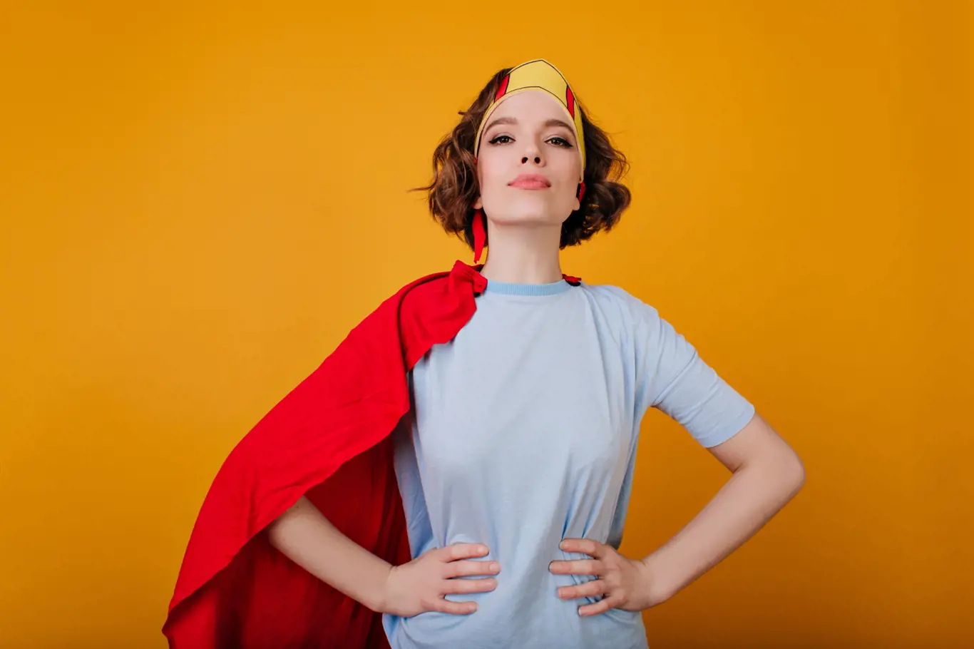 Confident curly girl in superhero attire posing on bright yellow background. Adorable lady with short hair wearing paper crown and red cloak.