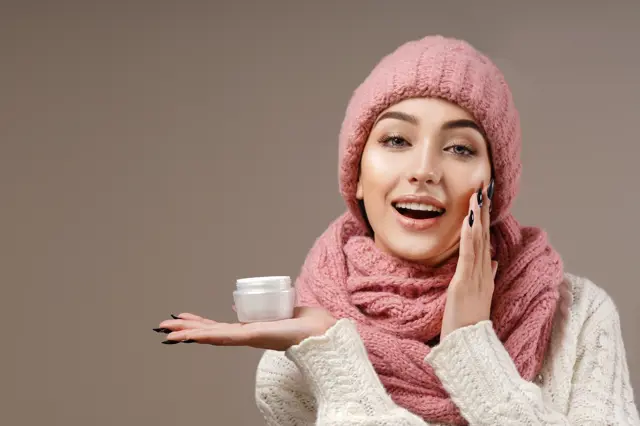 beauty face care portrait of attractive young caucasian woman in warm clothing studio shot toothy smiling winter applying cream